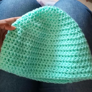 Puede incluir: Un gorro de ganchillo verde menta. El gorro está hecho a mano con un patrón de puntada texturizada. El gorro lo sostiene una persona. El gorro es de color liso y parece suave y cómodo.