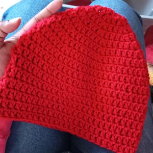 May include: A handmade, bright red crochet beanie hat. The hat is made with a tight stitch pattern, giving it a textured appearance. The hat is shown against a background of blue jeans.