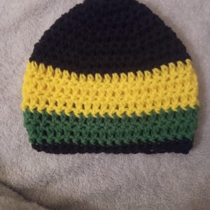 Puede incluir: Un gorro de ganchillo hecho a mano en negro, amarillo y verde. El gorro presenta un diseño a rayas con una parte superior negra, una sección media amarilla y una sección inferior verde. El gorro probablemente está hecho de hilo suave.