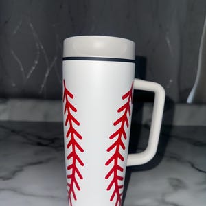 Puede incluir: Taza blanca con tem&aacute;tica de b&eacute;isbol con detalles de costuras rojas. La taza tiene un asa blanca y un borde gris. El texto "24oz" est&aacute; impreso en la parte inferior de la taza. Ideal para los fan&aacute;ticos del b&eacute;isbol.