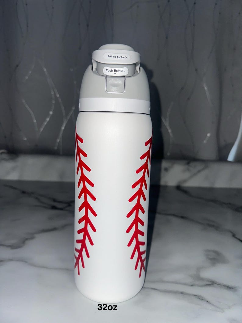 Puede incluir: Botella de agua blanca de 946 ml con dise&ntilde;o de costura de b&eacute;isbol roja. La botella tiene una tapa gris con un bot&oacute;n y las palabras "Lift to Unlock" y "Push Button".