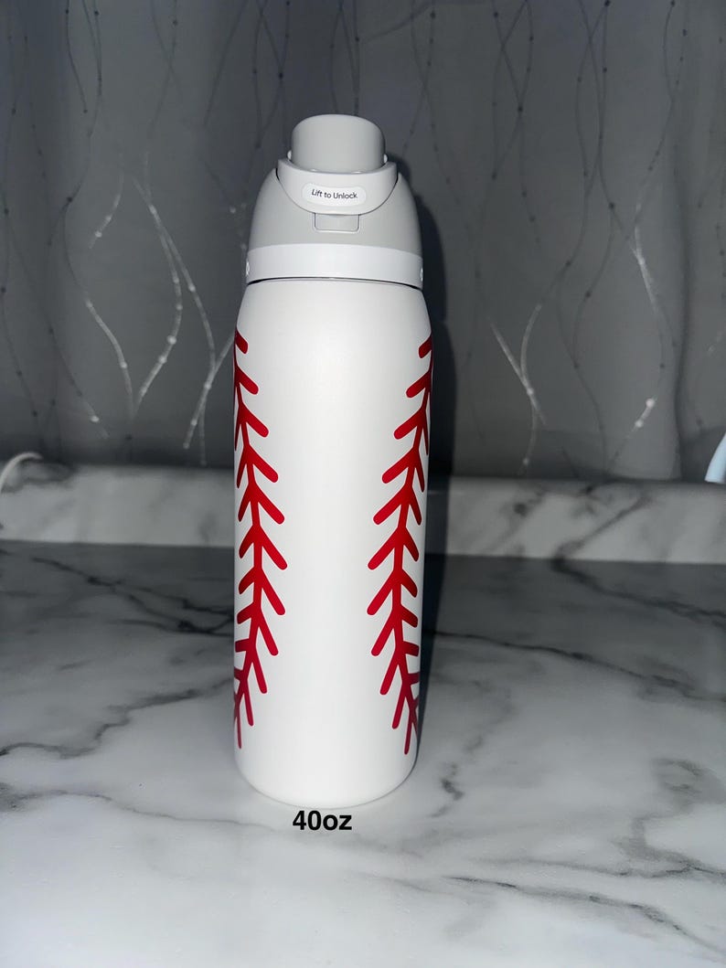 Puede incluir: Botella de agua blanca de 1,18 litros con dise&ntilde;o de costura de b&eacute;isbol roja. La botella tiene una tapa gris con un mecanismo de apertura de bot&oacute;n. El texto "Lift to Unlock" est&aacute; impreso en la tapa.
