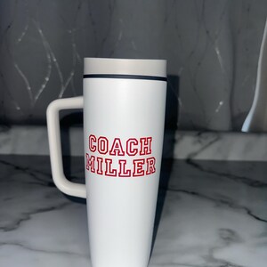 Puede incluir: Taza de viaje blanca con asa, con el texto "COACH MILLER" en letras rojas. La taza tiene una tapa gris y una banda negra. La parte inferior de la taza dice "24oz", que son aproximadamente 710 ml.