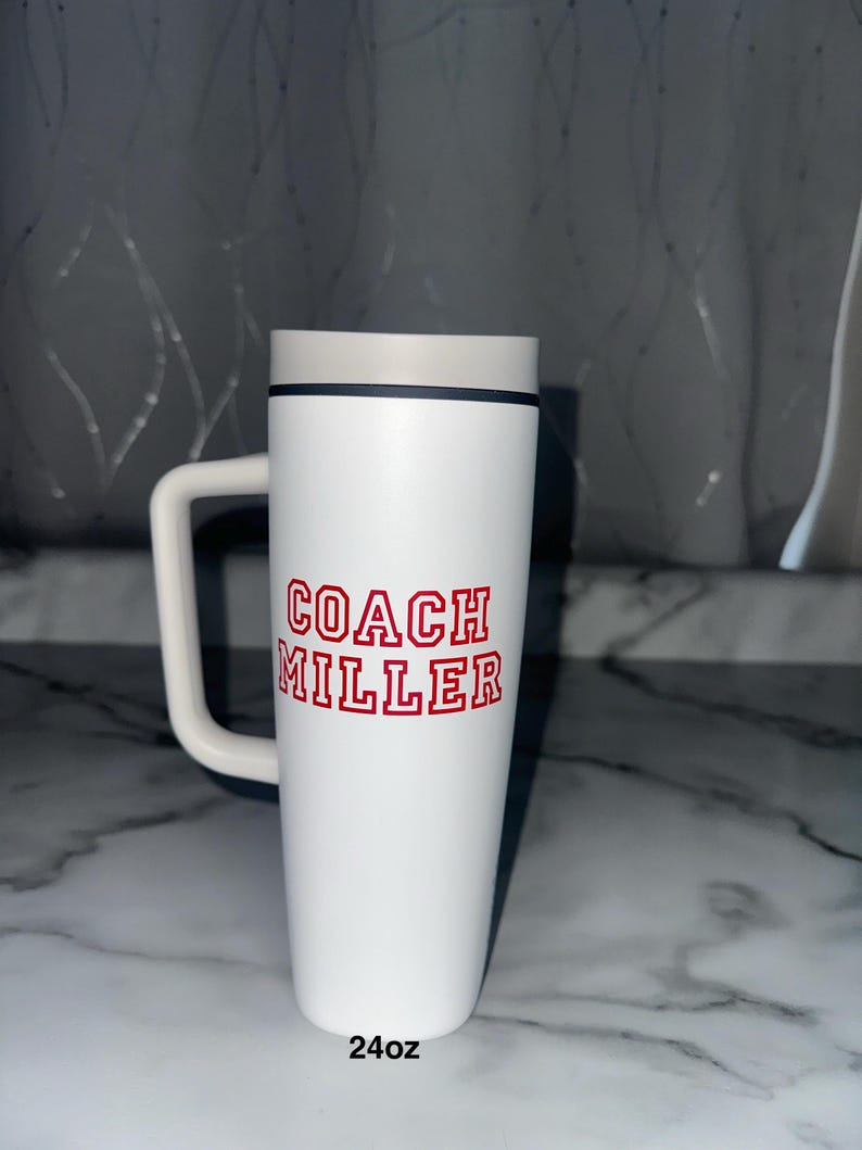 Puede incluir: Taza de viaje blanca con asa, con el texto "COACH MILLER" en letras rojas. La taza tiene una tapa gris y una banda negra. La parte inferior de la taza dice "24oz", que son aproximadamente 710 ml.
