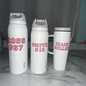 Puede incluir: Tres botellas de agua Owala blancas y una taza con texto rojo. Las botellas tienen tapas grises y la taza tiene un asa. Las botellas est&aacute;n etiquetadas "ROSS #67" y "SMITH #19". La taza est&aacute; etiquetada "COACH MILLER".