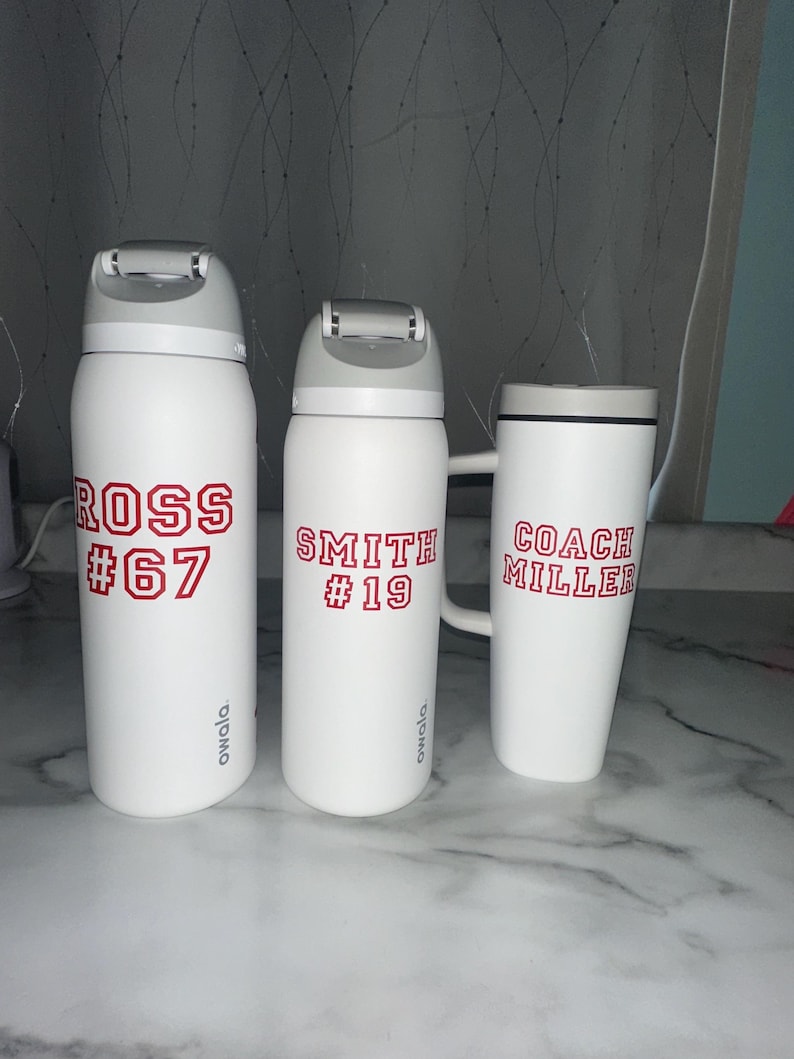 Puede incluir: Tres botellas de agua Owala blancas y una taza con texto rojo. Las botellas tienen tapas grises y la taza tiene un asa. Las botellas est&aacute;n etiquetadas "ROSS #67" y "SMITH #19". La taza est&aacute; etiquetada "COACH MILLER".