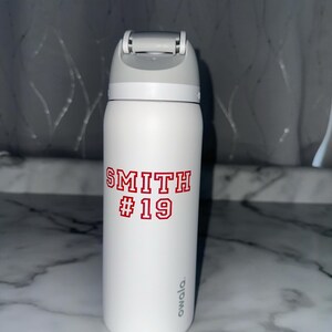 Puede incluir: Botella de agua aislada blanca con tapa gris. La botella presenta el nombre "SMITH" y el n&uacute;mero "19" en letras rojas. La botella es de 946 ml y tiene la marca "owala" impresa en el lateral.