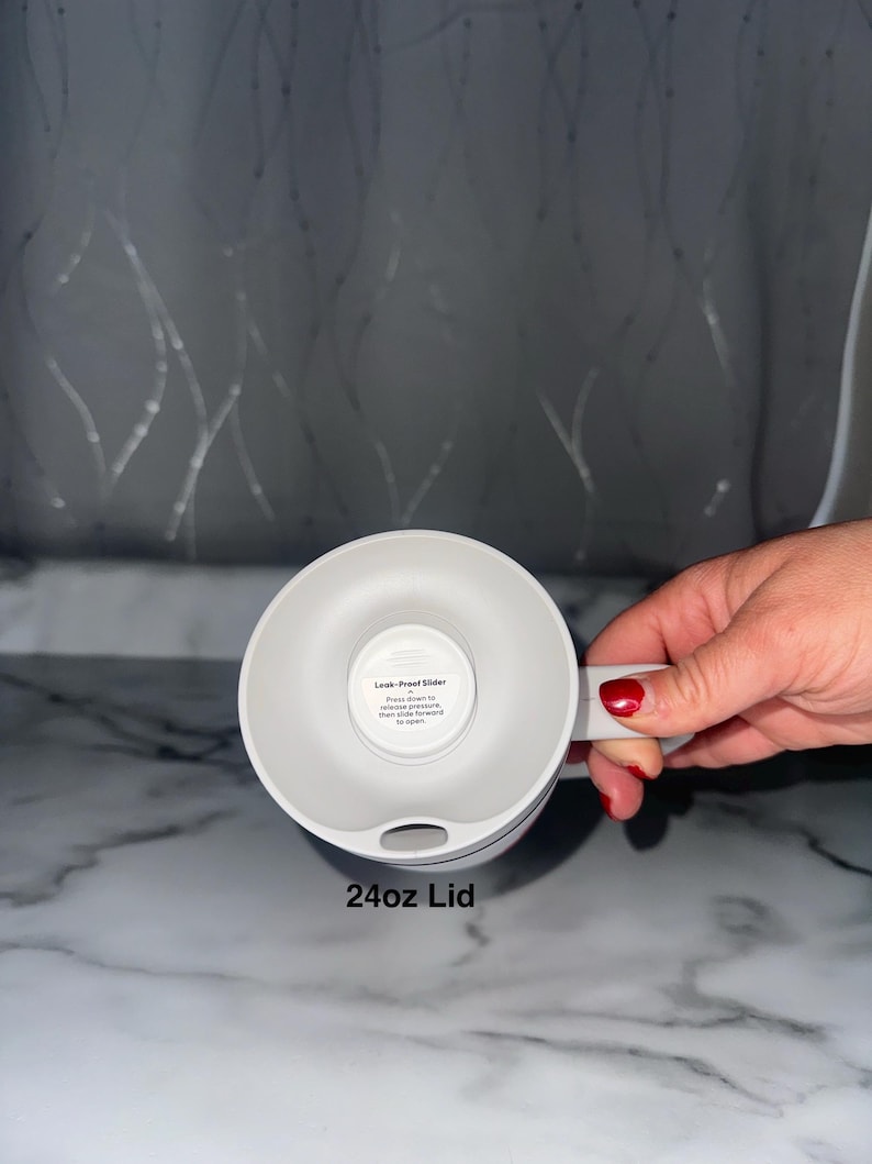 Puede incluir: Una tapa blanca de 24 oz para una taza, con un asa y un mecanismo deslizante a prueba de fugas. La tapa se muestra desde abajo, con el texto "24oz Lid" en la parte inferior. La tapa est&aacute; dise&ntilde;ada para mantener las bebidas seguras y evitar derrames.