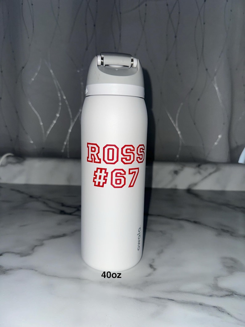 Puede incluir: Botella de agua aislada blanca con tapa abatible gris. La botella presenta el nombre "ROSS" y el n&uacute;mero "67" en letras rojas. La botella est&aacute; etiquetada como "40oz", que equivale a aproximadamente 1,18 litros.