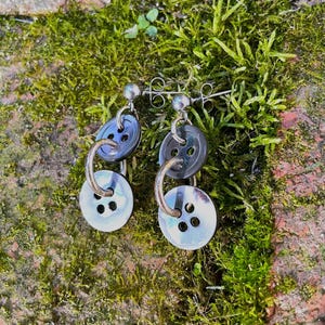 May include: A pair of handmade button earrings. Each earring features a dark gray button connected to a white button with a silver hoop. The earrings are set against a background of green moss and brick.