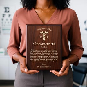 May include: A wooden plaque with the text "A Prayer For Optometrists" and a medical symbol. The plaque is held by a person. The text includes a prayer and the name "Dr. Jennifer Brown". The plaque is brown with gold lettering.