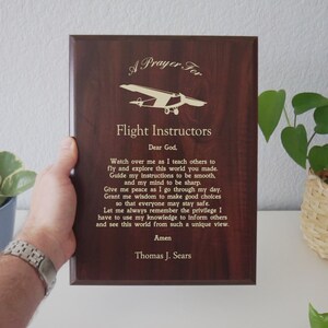 Pode incluir: Placa de madeira castanha escura com letras douradas, com uma oração para instrutores de voo e um gráfico de avião. O texto inclui uma oração e o nome Thomas J. Sears.