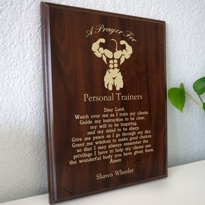 May include: A wooden plaque with a gold inscription that reads "A Prayer For Personal Trainers". The inscription includes a prayer for personal trainers, asking for guidance and wisdom in their work. The plaque is signed "Shawn Wheeler".