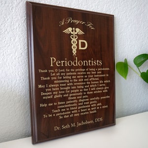 May include: A dark brown wooden plaque with gold lettering. The plaque has a prayer for periodontists, a medical symbol, and the text "A Prayer For Periodontists". The plaque is signed "Dr. Seth M. Jacobsen, DDS".
