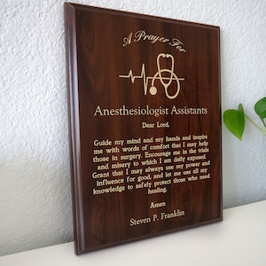May include: A brown wooden plaque with a gold inscription that reads "A Prayer For Anesthesiologist Assistants". The inscription includes a stethoscope and a heartbeat graphic.