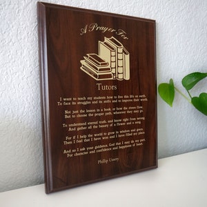 Op de afbeelding: Een houten plaquette met een gouden inscriptie die "Een gebed voor Tutoren" luidt. De inscriptie bevat een gedicht over lesgeven en het belang van karakter en zelfvertrouwen. De plaquette toont een stapel boeken in de linkerbovenhoek.