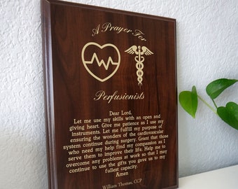Può includere: Una targa in legno con un'iscrizione dorata che recita "Una preghiera per i perfusionisti". La targa presenta un cuore dorato con una linea di battito cardiaco e un simbolo di caduceo dorato.