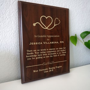 Op de afbeelding: Een bruine houten plaquette met een gouden inscriptie die luidt: "In Grateful Appreciation to Jessica Villabona, RN". De plaquette heeft een gouden stethoscoop en hartontwerp. De tekst gaat verder: "There are no words to express our deep felt thanks. Your caring, compassion, and medical knowledge have been invaluable to us. Thank you for guiding us into the field of nursing. West University Nursing Program Class 24-B."
