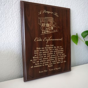 May include: A brown wooden plaque with a gold inscription that reads "A Prayer For Code Enforcement". The inscription includes a prayer for guidance and protection for code enforcement officers. The plaque is decorated with a simple line drawing of a neighborhood.