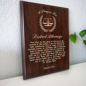 May include: A dark brown wooden plaque with gold lettering. The plaque has a prayer for district attorneys, a scale of justice, and the quote "St. Thomas More".