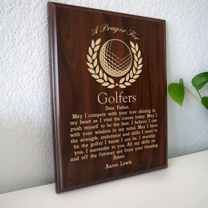 Op de afbeelding: Een bruine houten plaquette met een gouden golfbal en een lauwerkransontwerp. De plaquette heeft een gebed voor golfers erop gegraveerd, dat luidt: "Een gebed voor golfers Lieve Vader. Moge ik concurreren met uw liefde die schijnt in mijn hart terwijl ik vandaag de baan bezoek. Moge ik mezelf pushen om de beste te zijn. Ik geloof dat ik kan met uw wijsheid in mijn geest. Moge ik de kracht, uithoudingsvermogen en vaardigheden hebben die ik nodig heb om de golfer te zijn die ik weet dat ik kan zijn. Ik aanbid u, ik geef me over aan u. Al mijn vaardigheden op en naast de fairway komen van uw zegen. Amen Aaron Lewis"