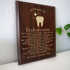 May include: A brown wooden plaque with gold lettering that reads "A Prayer For Endodontists". The plaque features a gold tooth with stars above it. The text includes a prayer for endodontists, thanking God for the opportunity to serve patients and asking for guidance and strength. The plaque is signed by Dr. Seth M. Jacobsen, DDM.