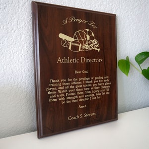 Op de afbeelding: Een houten plaquette met een gouden inscriptie die "Een gebed voor sportdirecteuren" luidt. De plaquette toont een gouden illustratie van een honkbalknuppel, een basketbal, een voetbal en een paar hardloopschoenen.