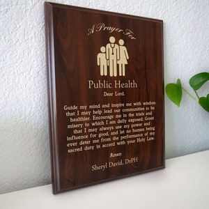 May include: A wooden plaque with a gold inscription that reads "A Prayer For Public Health." The inscription includes a prayer for guidance and strength in the field of public health. The plaque is signed "Sheryl David, DrPH."