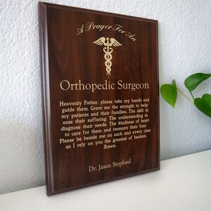 May include: A brown wooden plaque with gold lettering that reads "A Prayer For An Orthopedic Surgeon". The plaque features a gold medical symbol and a prayer for an orthopedic surgeon. The plaque is signed "Dr. Jason Stepford".
