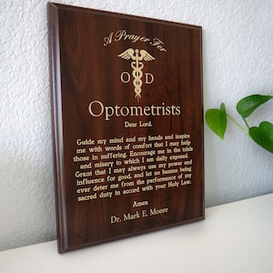 May include: A dark brown wooden plaque with a gold inscription. The plaque features a gold medical symbol with the letters "OD" and the text "A Prayer For Optometrists". The inscription includes a prayer for optometrists, ending with "Amen" and "Dr. Mark E. Moore".