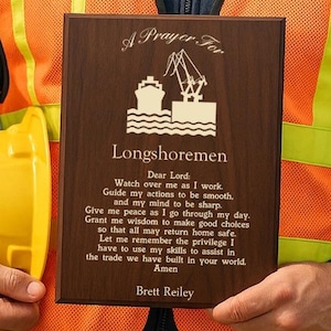 May include: A wooden plaque with the text "A Prayer For Longshoremen" and a ship graphic. The plaque is held by a person wearing an orange safety vest and a yellow hard hat. The text includes a prayer for longshoremen.