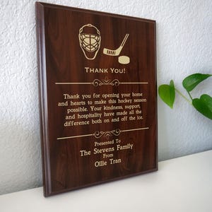 May include: A wooden plaque with a hockey theme. The plaque features a hockey goalie mask, a hockey stick, and a puck. The text reads "THANK YOU!" and a message of gratitude to the Stevens Family from Ollie Tran.