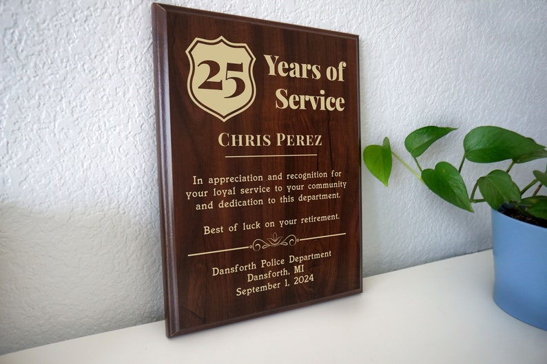 May include: A brown wooden plaque with gold lettering. The plaque reads "25 Years of Service" and "Chris Perez". The plaque also includes a gold shield with the number 25 inside. The plaque is a retirement award for a police officer.