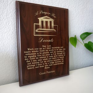 May include: A dark brown wooden plaque with gold lettering and a building graphic. The text reads "A Prayer For Docents" and includes a prayer by Grant Peterson. The plaque is displayed on a white surface.