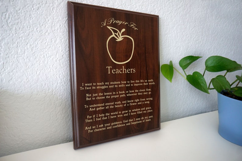May include: A wooden plaque with a dark brown finish, featuring the words "A Prayer For Teachers" and an apple graphic. The plaque includes an engraved poem. A potted plant is visible in the background.