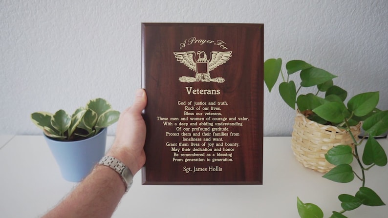 Puede incluir: Una placa de madera con un &aacute;guila dorada y el texto "A Prayer For Veterans" y un poema agradeciendo a los veteranos por su servicio. La placa est&aacute; sobre una superficie blanca con dos plantas en maceta.