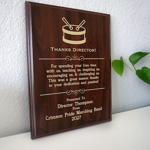 May include: A rectangular wooden plaque with a dark brown finish. The plaque features a drum and drumsticks graphic and the text "THANKS DIRECTOR!" along with a personalized message. The text includes the name "Director Thompson" and "Crimson Pride Marching Band 2027".