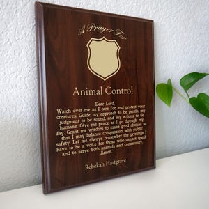 May include: A dark brown wooden plaque with gold lettering. The text reads "A Prayer For Animal Control" above a shield emblem and a prayer. The name Rebekah Hartgrave is at the bottom of the plaque.