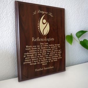 Op de afbeelding: Een donkerbruine houten plaquette met gouden letters. De tekst luidt "A Prayer For Reflexologists" en een gebed. De plaquette wordt gepresenteerd op een wit oppervlak.