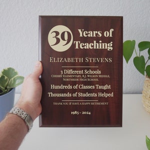 May include: A dark brown wooden plaque with gold lettering. The plaque reads "Years of Teaching" with the number "39" in a circle. The plaque also includes the name "Elizabeth Stevens", the text "3 Different Schools", "Hundreds of Classes Taught", "Thousands of Students Helped", "Thank You & Have a Happy Retirement", and the dates "1985-2024".