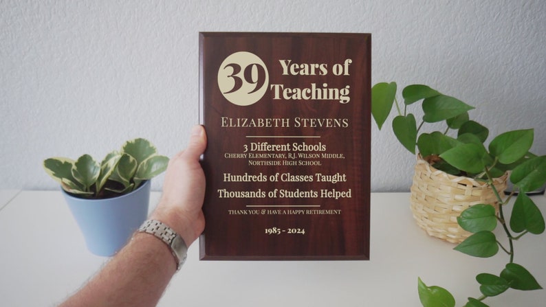 May include: A dark brown wooden plaque with gold lettering. The plaque reads "Years of Teaching" with the number "39" in a circle. The plaque also includes the name "Elizabeth Stevens", the text "3 Different Schools", "Hundreds of Classes Taught", "Thousands of Students Helped", "Thank You & Have a Happy Retirement", and the dates "1985-2024".