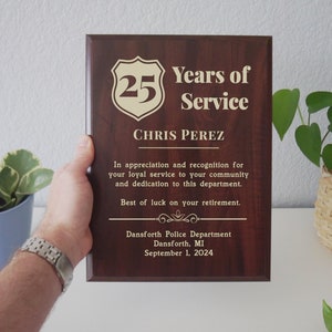 May include: A brown wooden plaque with gold lettering. The plaque reads "25 Years of Service" and "Chris Perez". The plaque also includes the text "In appreciation and recognition for your loyal service to your community and dedication to this department. Best of luck on your retirement." The plaque also includes the text "Dansforth Police Department, Dansforth, MI, September 1, 2024."