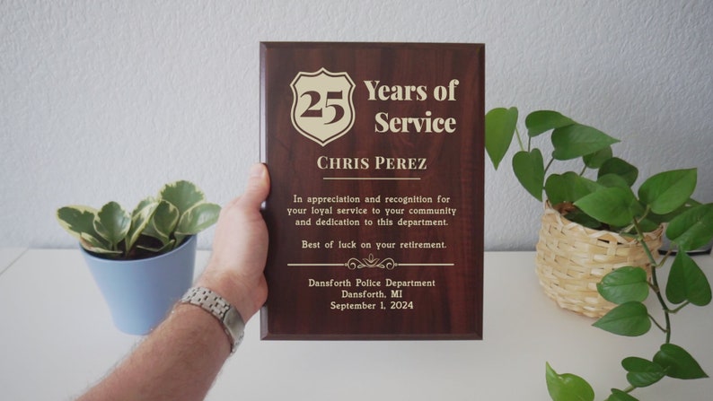 May include: A brown wooden plaque with gold lettering. The plaque reads "25 Years of Service" and "Chris Perez". The plaque also includes the text "In appreciation and recognition for your loyal service to your community and dedication to this department. Best of luck on your retirement." The plaque also includes the text "Dansforth Police Department, Dansforth, MI, September 1, 2024."