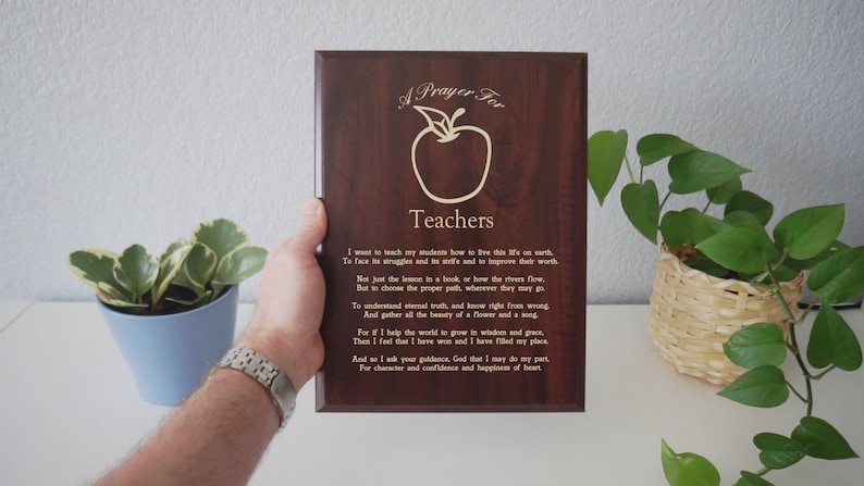 May include: A wooden plaque with the words "A Prayer For Teachers" and an apple illustration. The plaque has a dark wood grain finish and includes a prayer for teachers. The plaque is held by a person.
