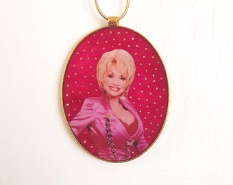 May include: A gold-framed oval keychain with a pink background and white polka dots. A photo of Dolly Parton is in the center of the keychain.