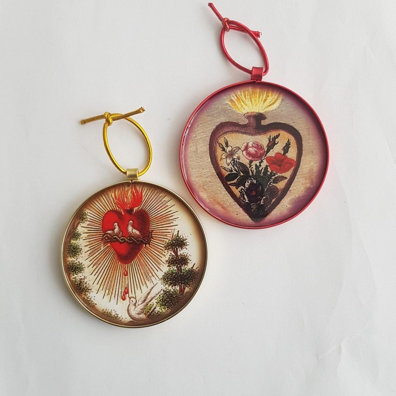 May include: Two round ornaments with religious imagery. One features a red heart with doves, flames, and rays, set in a gold frame. The other has a heart-shaped vase with flowers, in a red frame. Both have hanging loops.