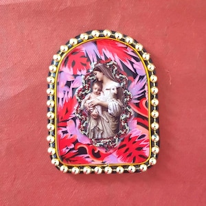 May include: A religious icon featuring a woman holding a child and a lamb, set against a floral background. The image is framed with a black border, accented with gold-colored beads. The overall color scheme includes red, pink, and gold.