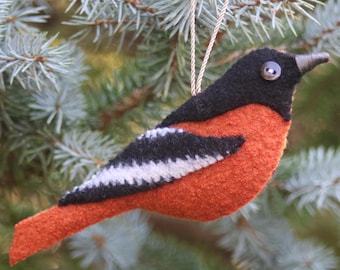 May include: A felt bird ornament featuring a black head, a bright orange body, and black wings with a white stripe. The bird has a small button eye and a pointed beak, and it is suspended by a thin silver cord for hanging as a decorative item.