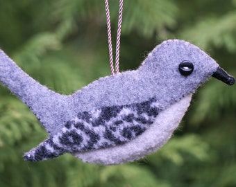 May include: A felt bird ornament in shades of light and dark gray. The bird features a black button eye, a small black beak, and a patterned wing with dark gray swirls. It hangs from a thin, brown and white twisted cord.