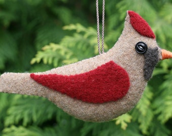 May include: A felt bird ornament in shades of tan and red, featuring a dark gray face patch, a small orange beak, and a black button eye. The bird has a red crest on its head and a red wing patch, suspended by a thin, braided silver cord.